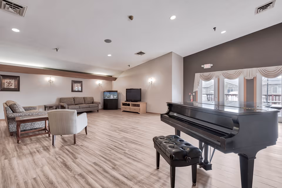 A spacious living room area in a senior living facility featuring a black grand piano with a cushioned bench in the foreground. The room has light wood flooring, beige walls, and a seating area with two sofas and an armchair arranged around a small table. A flat-screen TV sits on a wooden stand next to a fish tank. Large windows with white curtains allow natural light to enter the room.