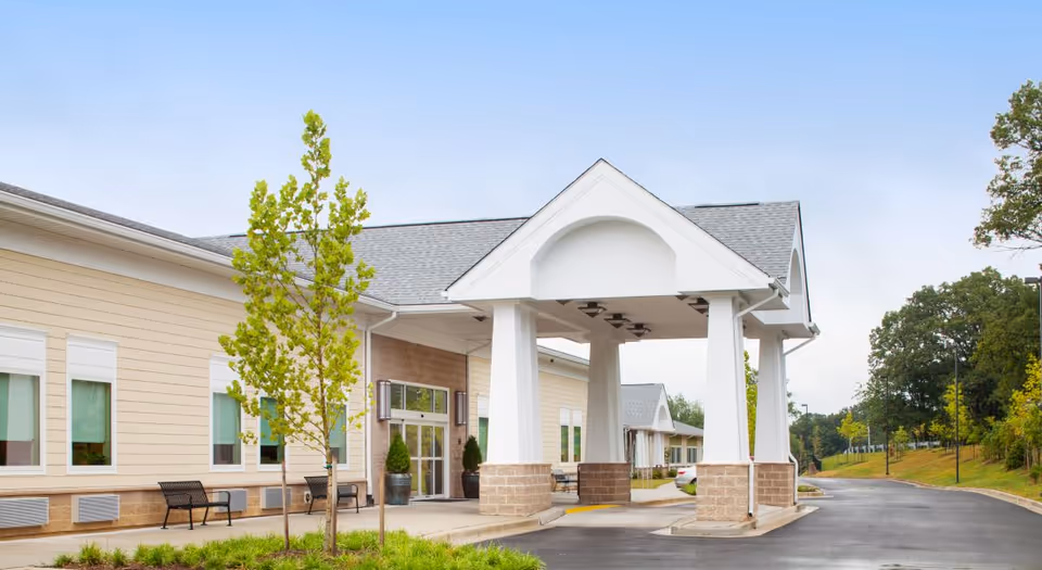 Exterior view of a single-story building with beige siding and white trim, featuring a covered entrance supported by white columns with stone bases. There are benches and small trees near the entrance, and a paved driveway leading up to the building. The sky is clear and blue.