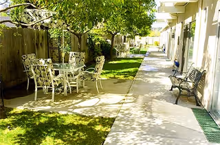 Sunny outdoor courtyard with a patio table and chairs under a tree alongside a walkway and benches outside facility rooms.