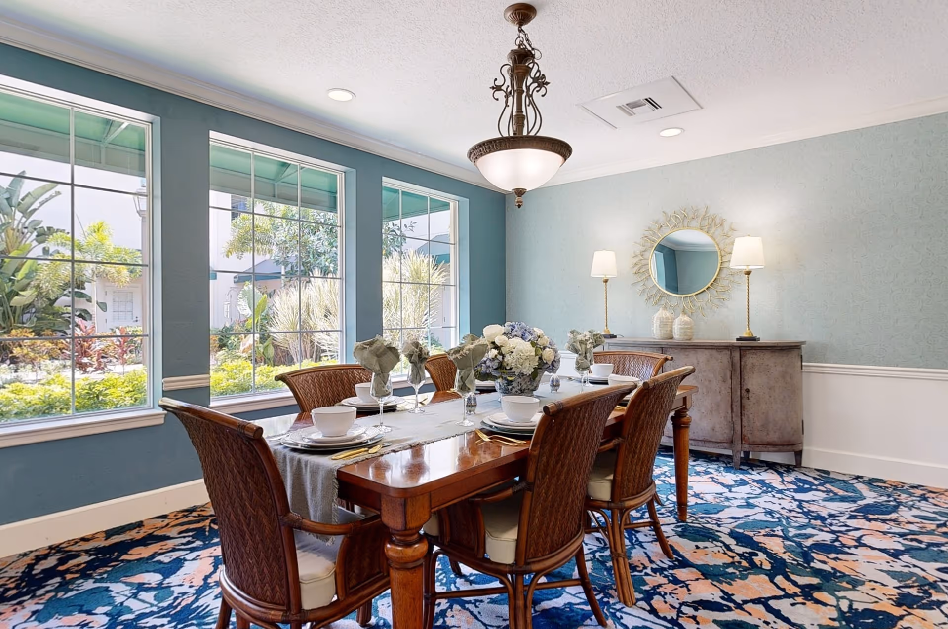 A dining room with a wooden table set for six people, featuring white bowls, plates, and glasses with napkins. The room has large windows showing greenery outside, blue walls, a patterned carpet, a chandelier, and a sideboard with two lamps and a decorative round mirror above it.