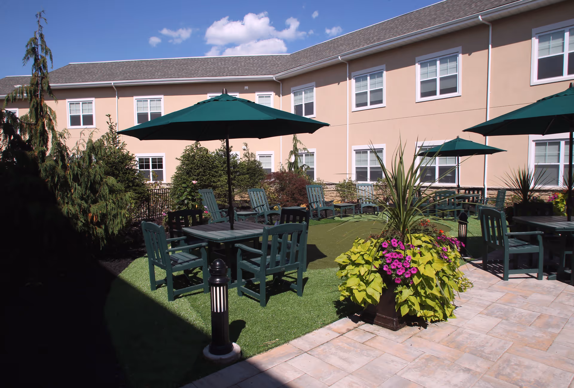 Outdoor patio area at Brandywine Livingston by Monarch featuring green tables and chairs with large green umbrellas, surrounded by plants and flowers, with a beige two-story building in the background under a blue sky with some clouds.