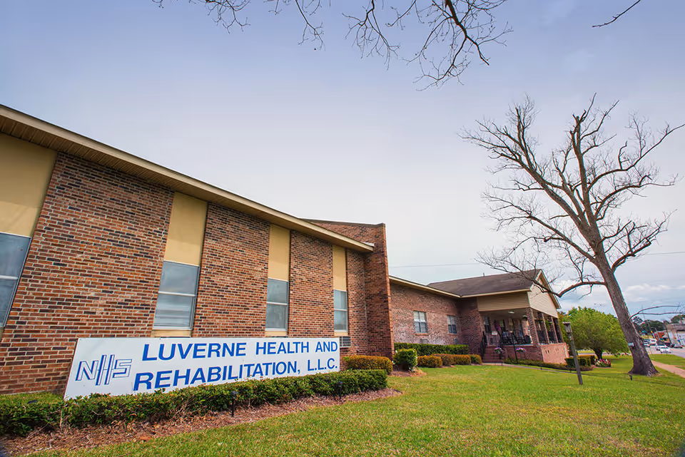 Exterior view of Luverne Health and Rehabilitation, LLC building with a brick facade, several windows, a lawn with bushes, and a large leafless tree in the foreground under a clear sky.