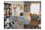 A cozy bedroom with a single bed covered in a floral bedspread, a cushioned armchair with a plaid blanket and stuffed toys, framed pictures on the walls, a window with lace curtains, and a radiator underneath the window.