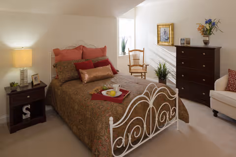 A cozy bedroom featuring a decorative white metal bed with patterned bedding, a nightstand with lamp, a dresser, and a chair by the window.