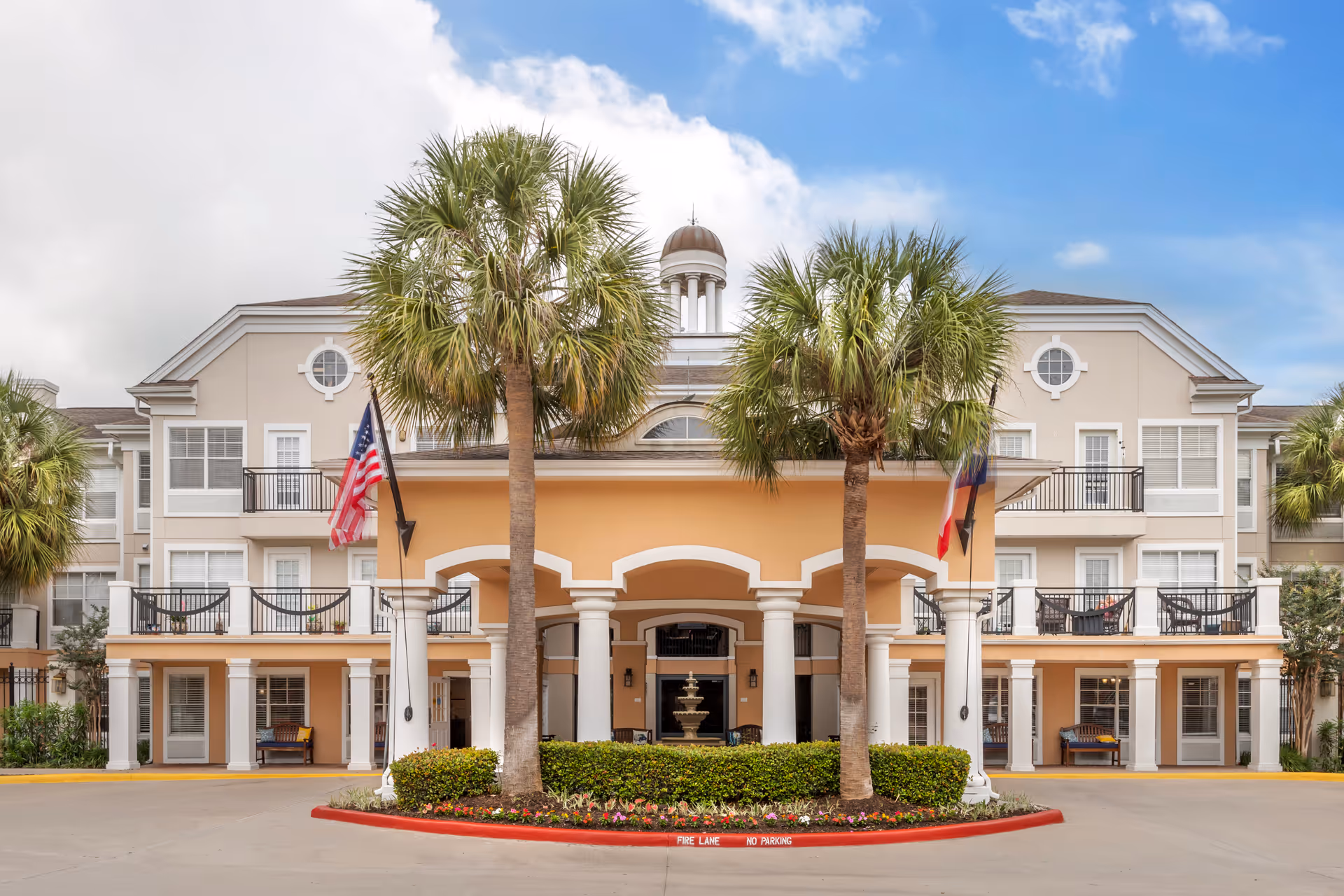 Front entrance of a multi-story senior living building with palm trees, columns, flags, balconies, and a fountain.