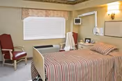 A hospital-style bedroom with a single bed covered in a striped bedspread, a red cushioned chair, a window with closed blinds and a floral valance, a bedside table with a lamp and framed photos, and medical equipment mounted on the wall.