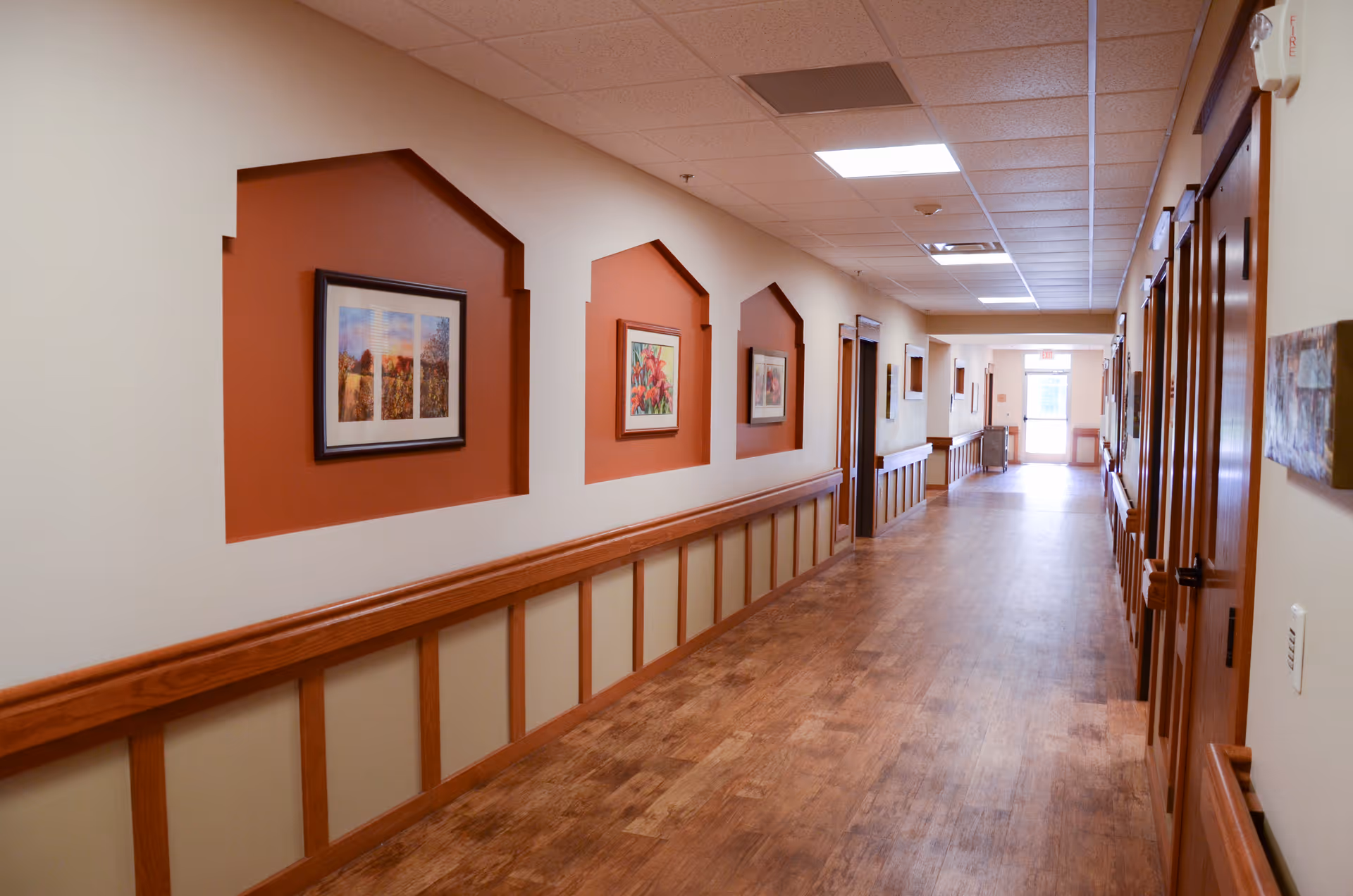Long interior hallway with wood flooring, framed artwork and wooden handrails leading to doors and an exit.