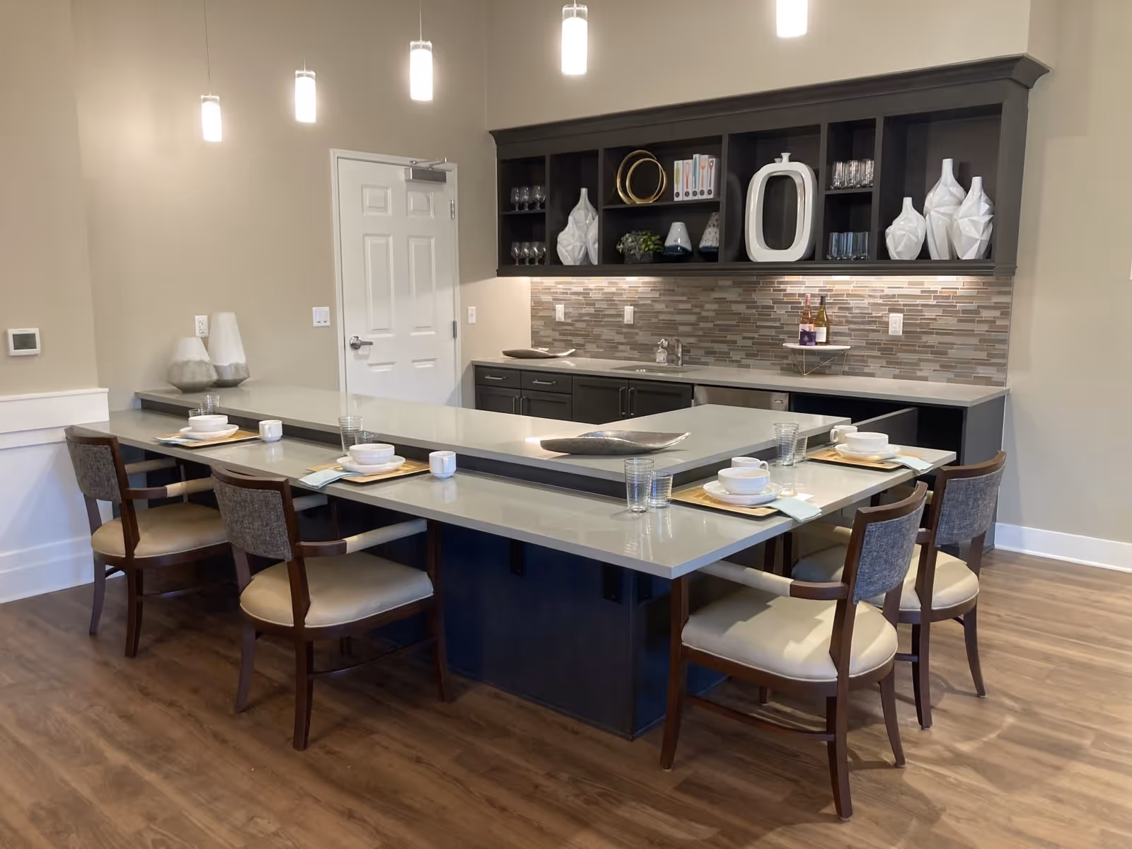 A modern dining area with a U-shaped counter and six chairs arranged around it. The counter is set with placemats, bowls, cups, and glasses. Above the counter, there are pendant lights hanging from the ceiling. Behind the counter, there are dark cabinets with decorative vases, glassware, and bottles of wine on the countertop. The floor is wooden, and the walls are painted beige.