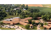 Aerial view of Leonardville Nursing Home facility surrounded by trees and farmland, showing multiple connected buildings with brown roofs and parking areas with cars.