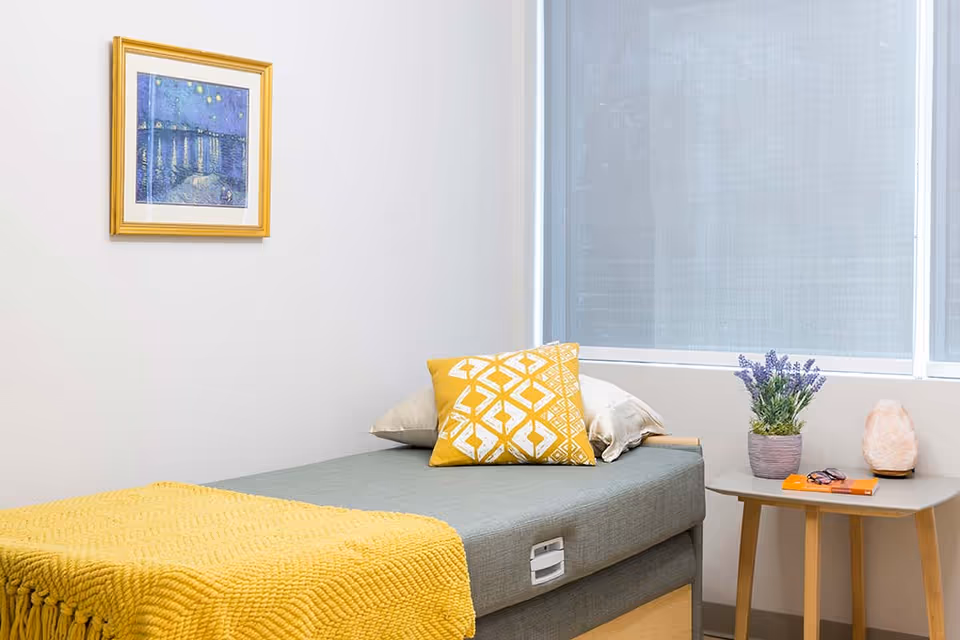 Small bright bedroom with a single bed dressed in gray and yellow linens, a side table holding a plant and lamp, and framed artwork on the wall.