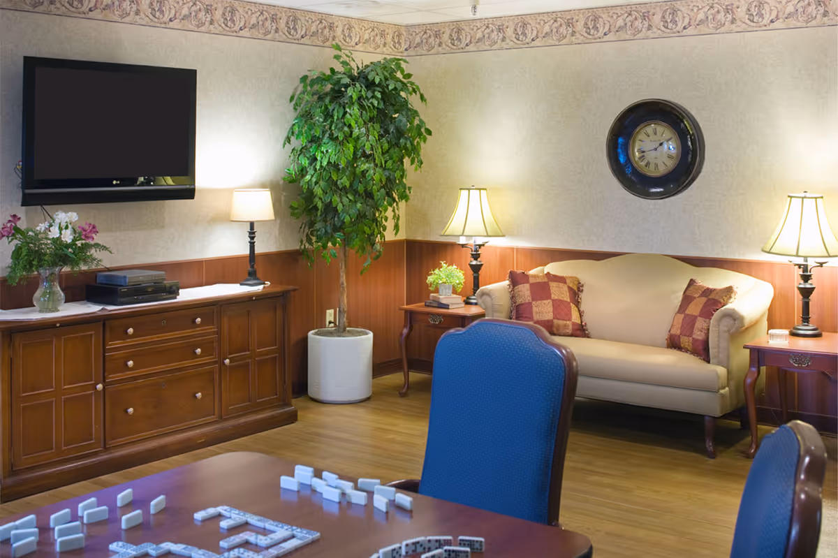 A cozy living room area with a beige couch adorned with two patterned pillows, two side tables each with a lamp, a large wall clock, a tall potted plant, a wooden cabinet with a flat-screen TV mounted above it, and a table in the foreground with dominoes set up for a game.