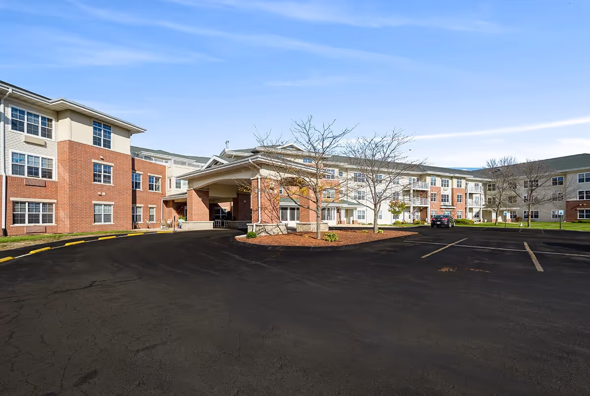 Exterior view of a multi-story senior living facility building with a covered entrance, parking lot, and some leafless trees under a clear blue sky.