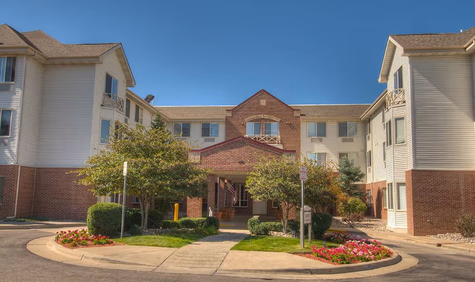 Exterior front view of Caley Ridge Assisted Living facility showing a three-story building with brick and light-colored siding, a covered entrance, landscaped greenery, trees, and flower beds under a clear blue sky.