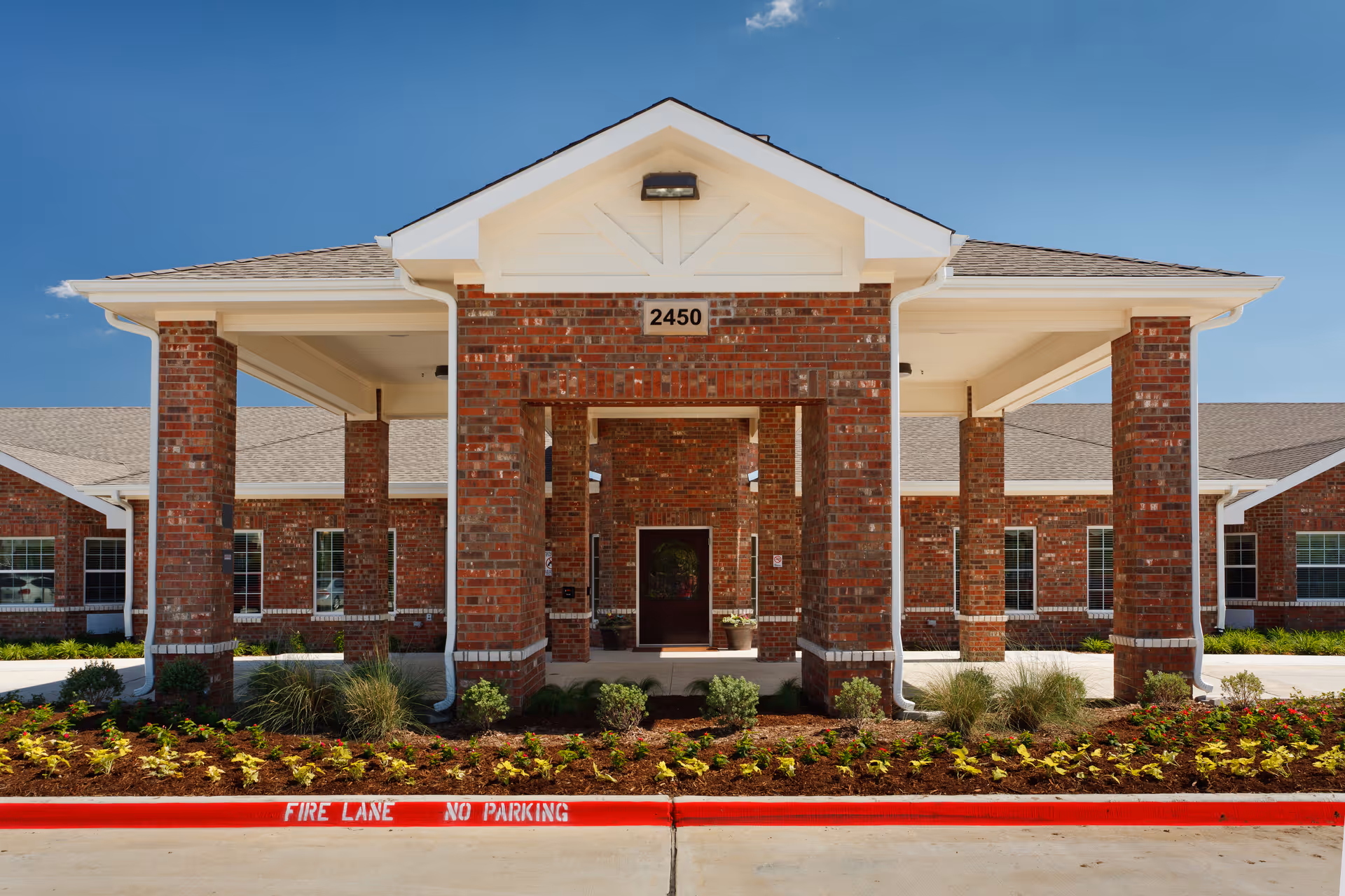 Front entrance of a brick senior living facility with a covered porte-cochere, columns, address number 2450, and landscaped flowerbeds.