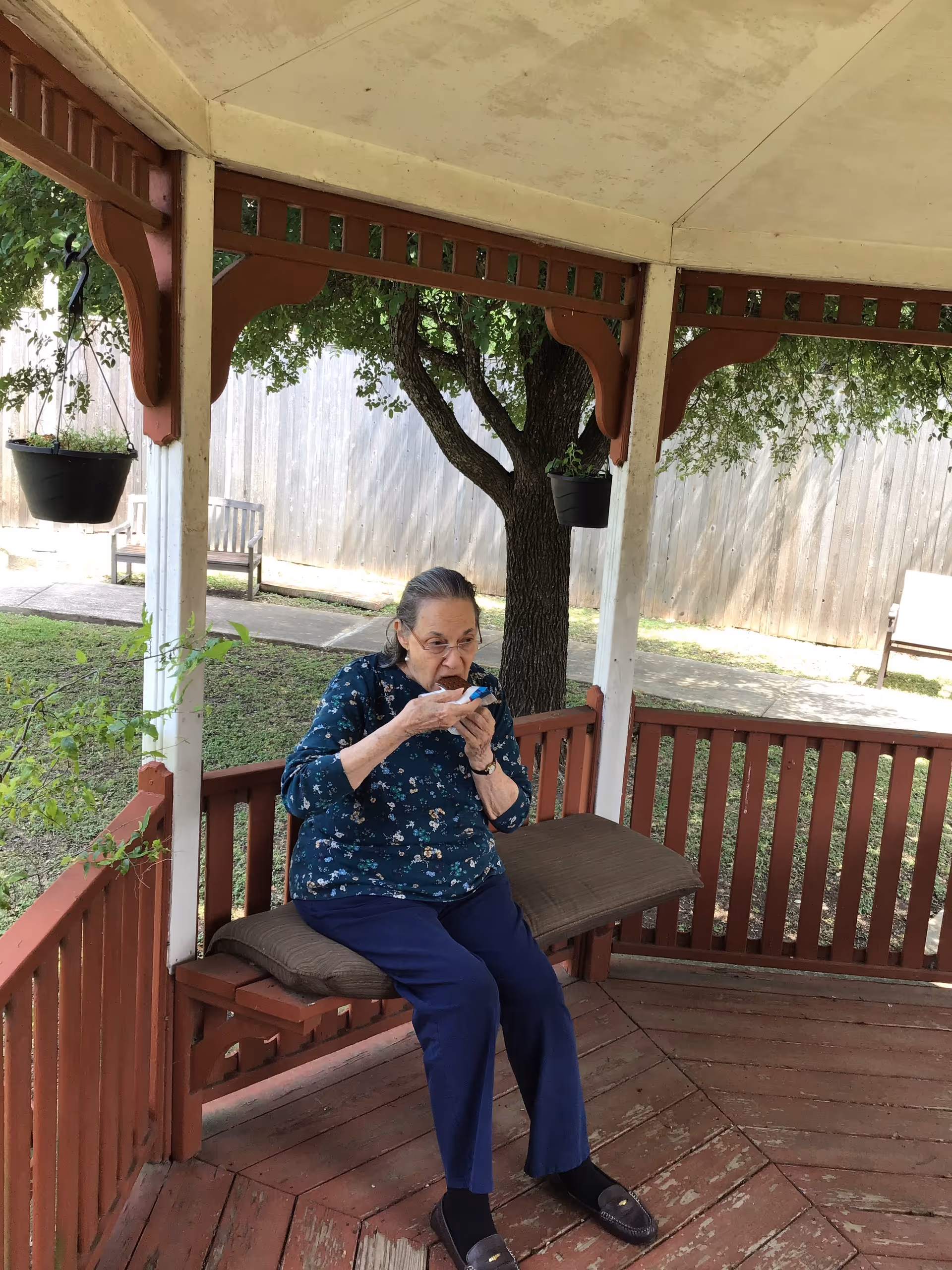An elderly woman sitting on a cushioned bench inside a wooden gazebo, eating a cupcake. The gazebo is painted red and white, surrounded by greenery and trees, with a wooden fence and benches visible in the background.