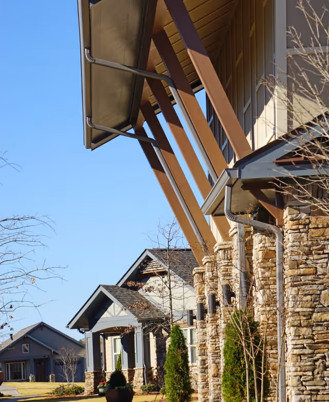 Close-up of a senior living facility's stone-clad front with large angled wooden support beams and neighboring units under a clear blue sky.