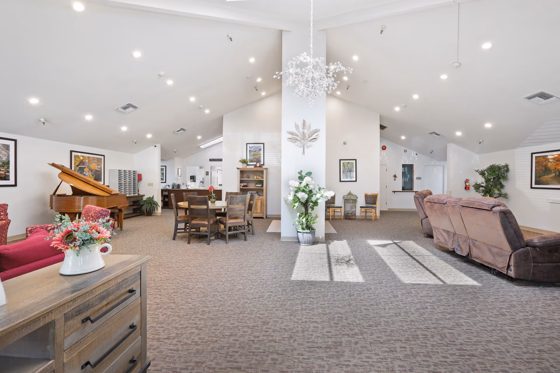 A spacious and well-lit common area in Almond Grove Assisted Living featuring a grand piano, a round wooden table with chairs, comfortable recliner sofas, decorative plants, framed artwork on the walls, and a chandelier hanging from the ceiling.