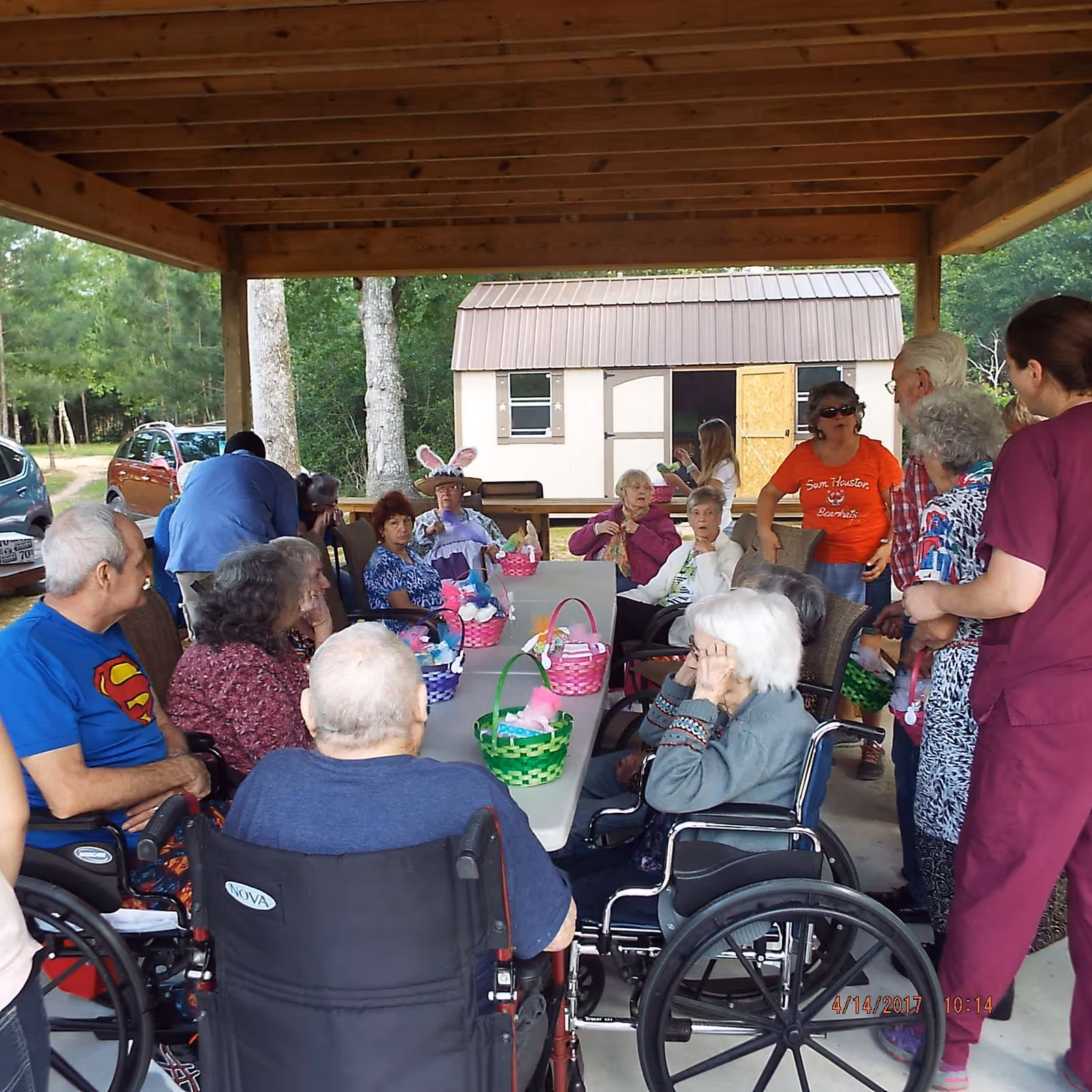 A group of elderly people, some in wheelchairs, gathered under a wooden pavilion around a long table with colorful Easter baskets. Several caregivers and visitors are standing nearby, and a small shed is visible in the background surrounded by trees.