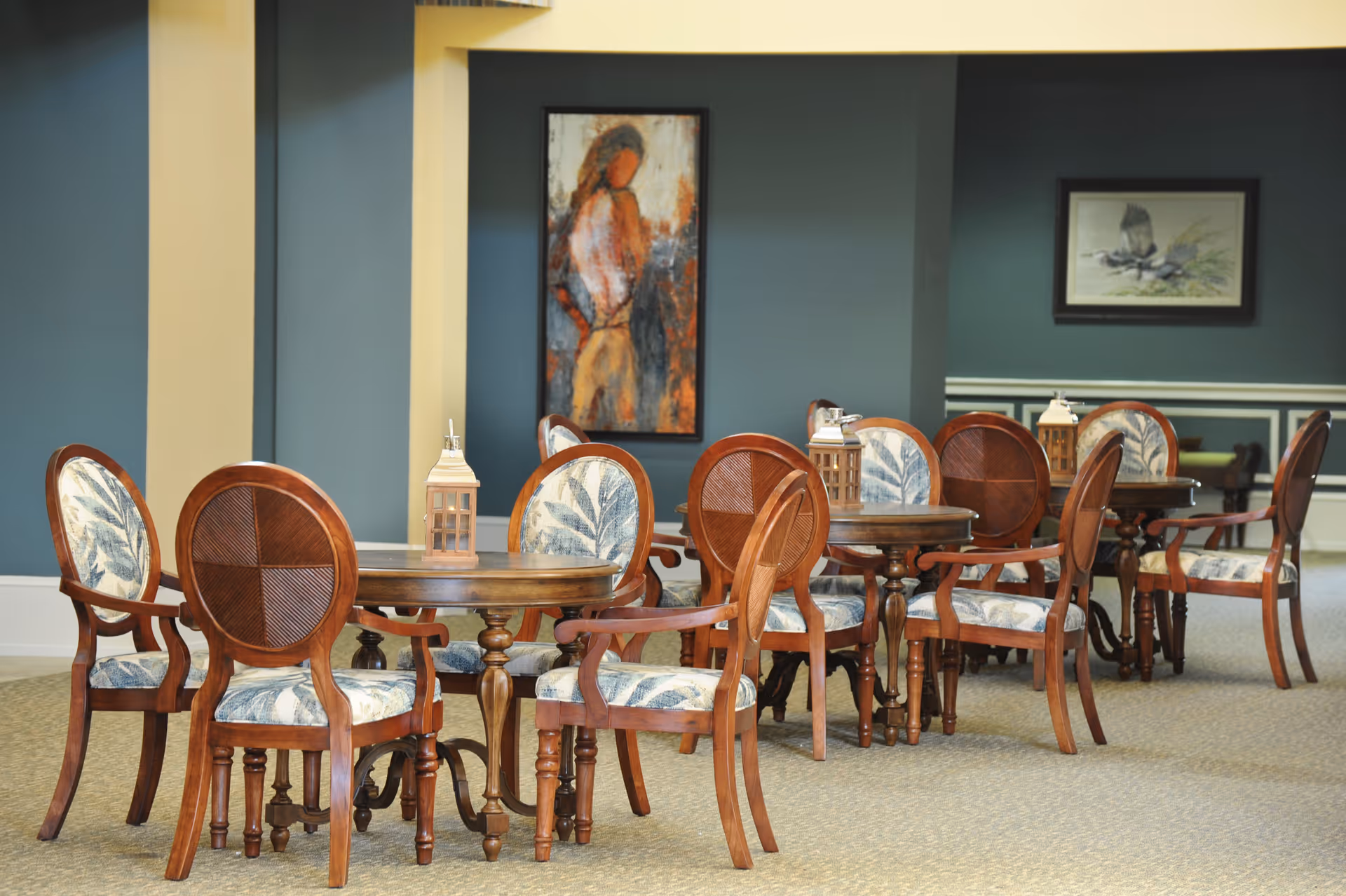 A dining area with several round wooden tables and matching chairs upholstered with a leaf-patterned fabric. Each table has a decorative lantern on it. The walls are painted blue with white trim, and two framed artworks hang on the walls in the background.