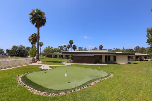 A small putting green and well-kept lawn with palm trees in front of a low single-story building under a clear blue sky.