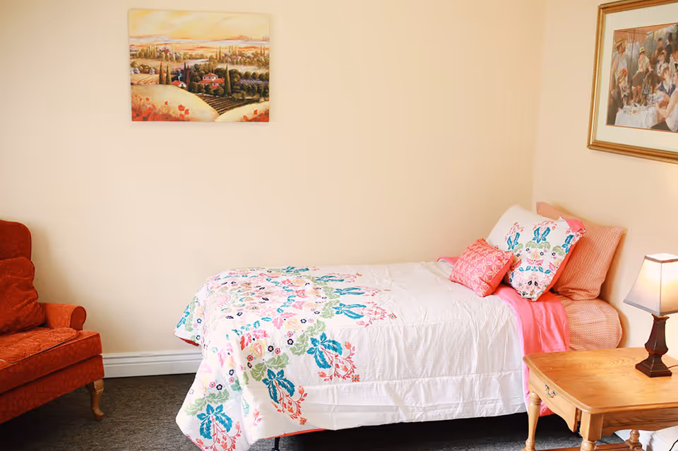 Single bedroom with a floral quilted bed, wooden nightstand and lamp, red upholstered chair, and framed wall art.