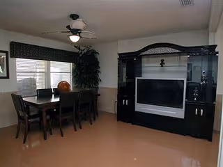 Interior view of a room with a dining table and six chairs near a window with blinds and a valance. A ceiling fan with a light is above the table. To the right, there is a large black entertainment center with a flat-screen TV and decorative items. A tall potted plant is placed in the corner between the dining area and the entertainment center.