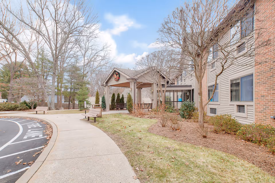 Entrance of a senior living facility with a covered porte-cochère, walkway, benches, and landscaped grounds beside the building.