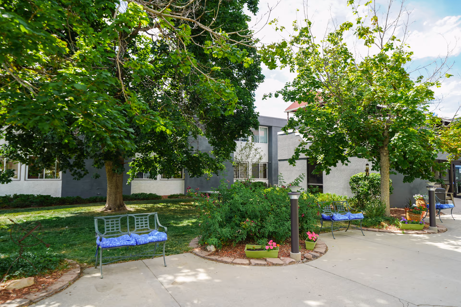 Outdoor courtyard area at Cherrelyn Health Care Center with green trees, bushes, and benches with blue cushions. The building exterior is visible in the background under a partly cloudy sky.