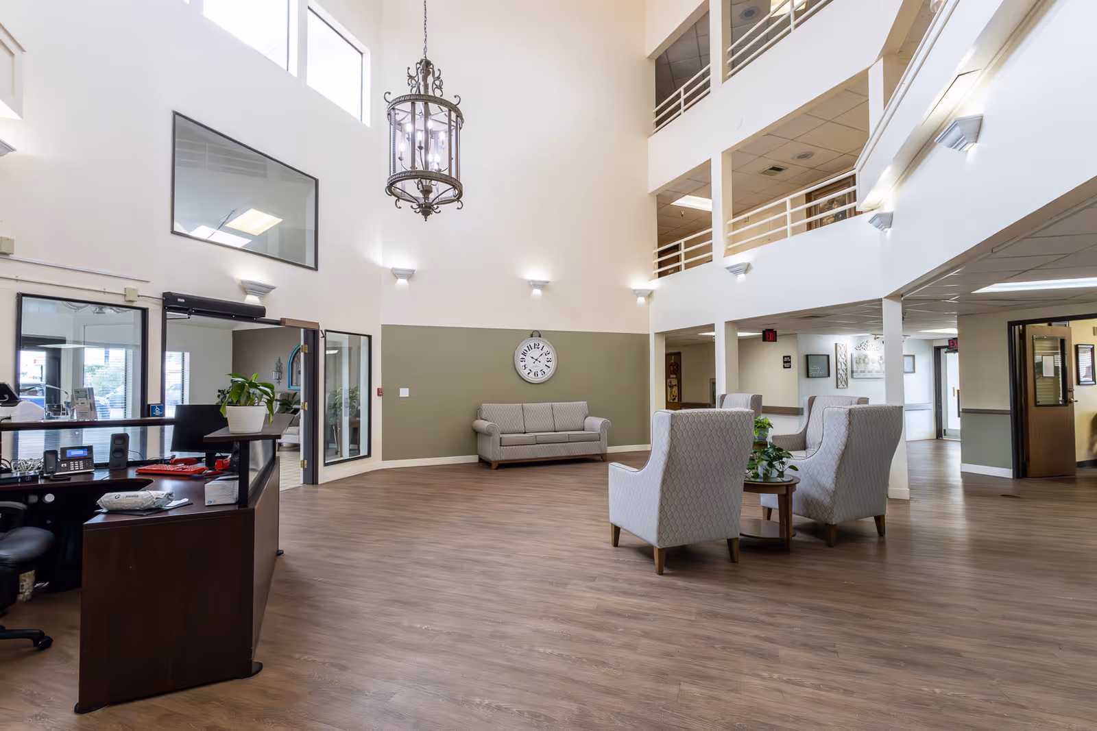 Spacious and well-lit lobby area of a senior living facility with high ceilings, a large hanging chandelier, a wall clock, a reception desk with office equipment, and seating arrangements including a sofa and armchairs around a small table with a plant.