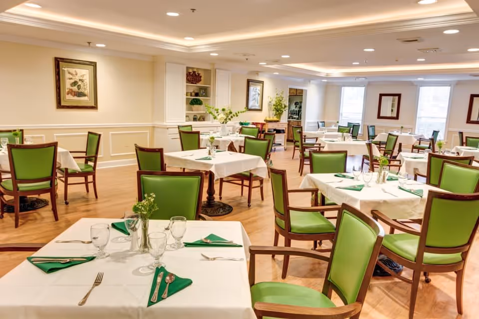 Dining room with neatly set tables, white tablecloths, and green-upholstered chairs.