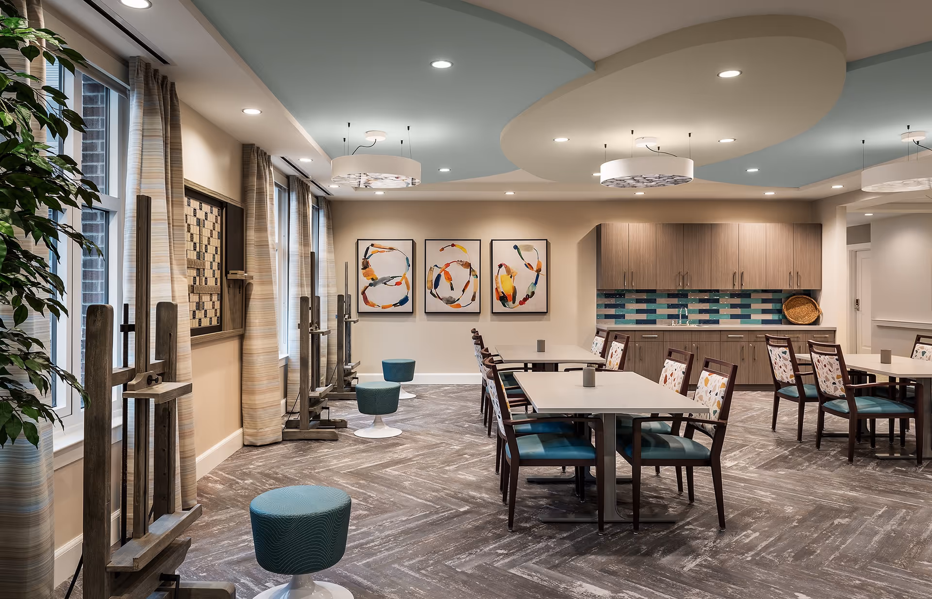 A well-lit, modern activity room with tables and chairs arranged for group seating. The room features a decorative ceiling with recessed lighting and hanging fixtures, a wall with colorful abstract artwork, and a cabinet area with a tiled backsplash. Several wooden easels and small stools are positioned near large windows with striped curtains, suggesting an art or craft space.