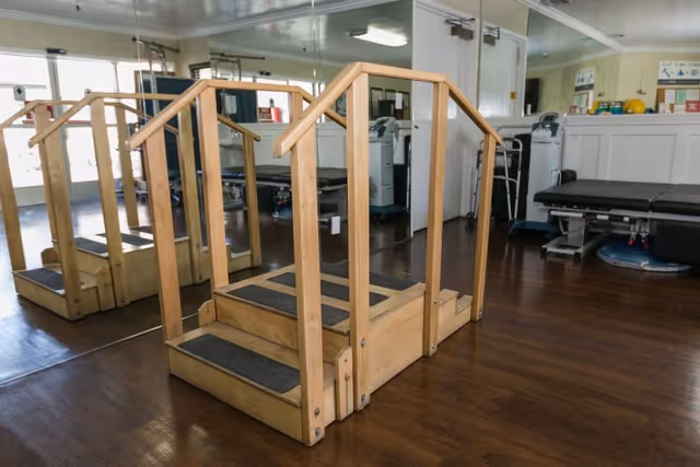 Physical therapy room with wooden parallel bars and steps for rehabilitation exercises, mirrored wall, medical equipment, and treatment tables in the background.