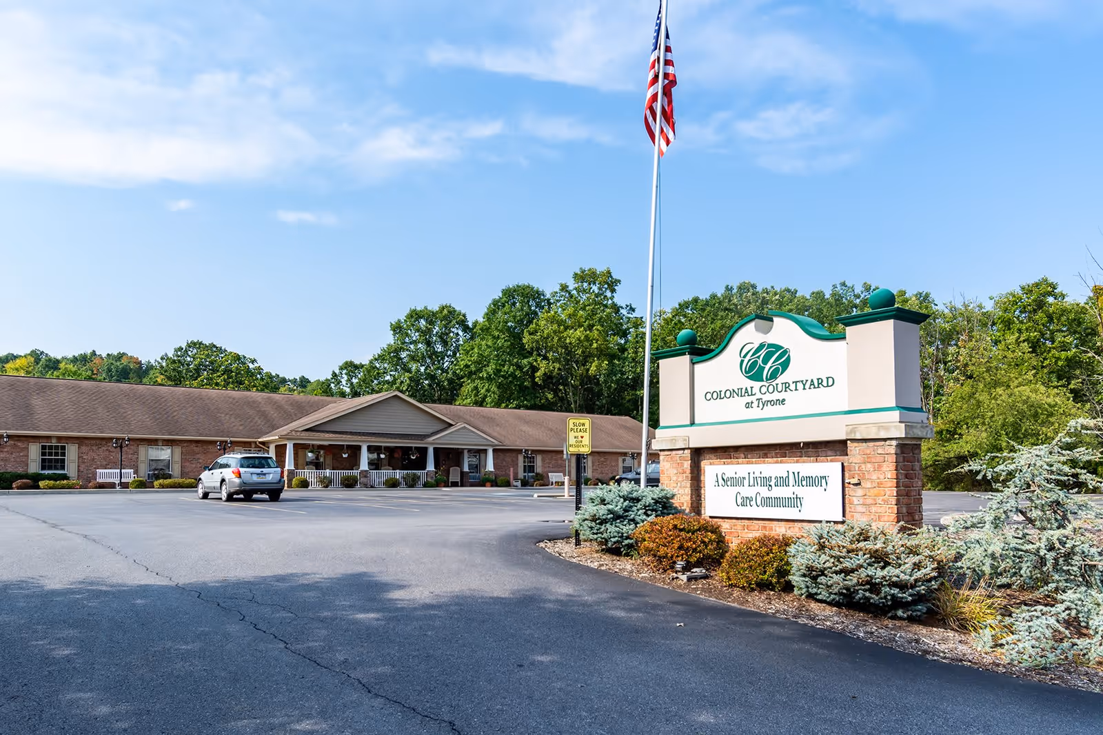 Exterior view of Colonial Courtyard at Tyrone, a senior living and memory care community. The image shows a single-story brick building with a covered entrance, a parking lot with a car, an American flag on a flagpole, and a large sign with the facility's name and description surrounded by landscaping and trees under a partly cloudy sky.