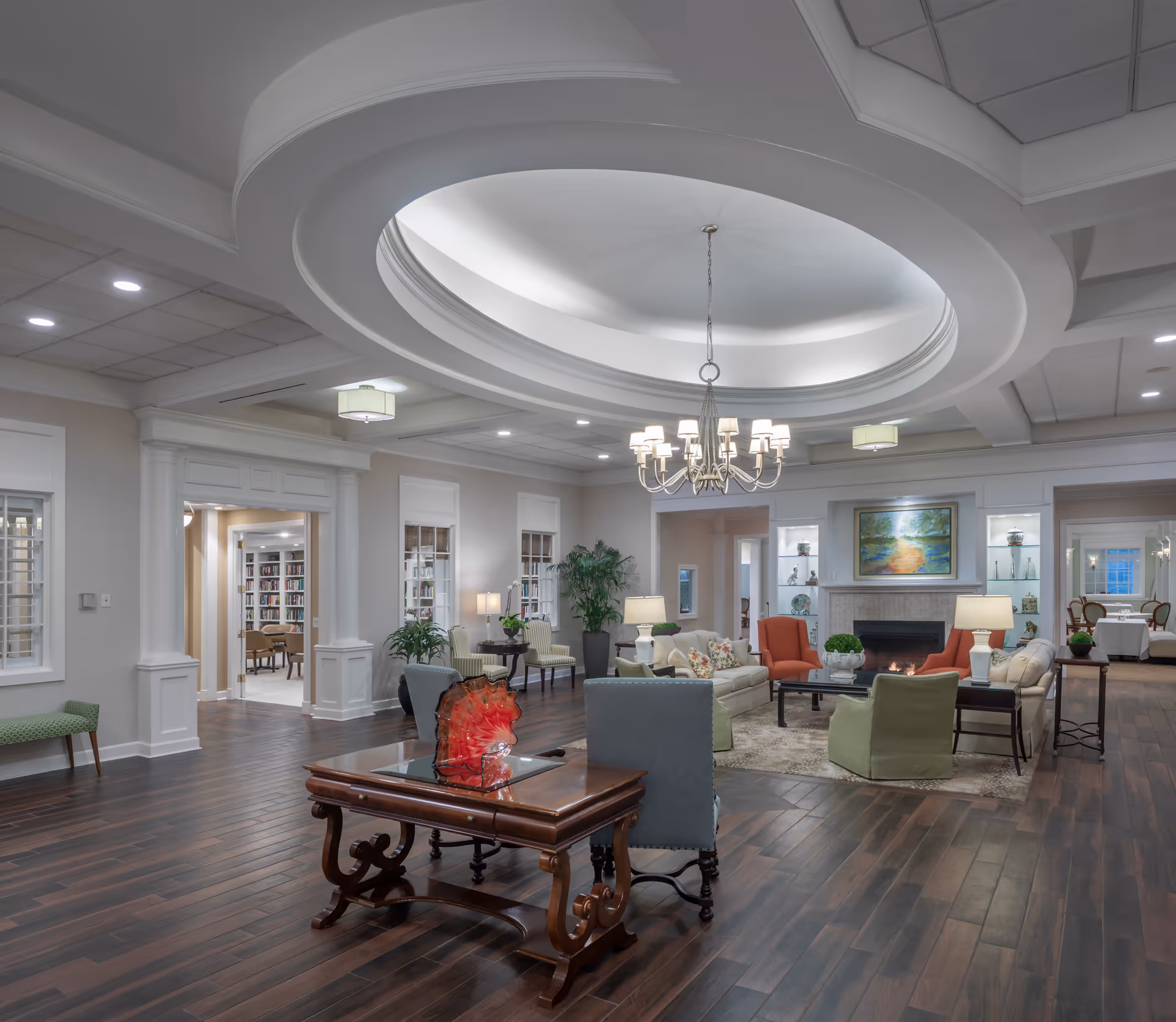 Spacious, elegantly furnished common lounge with seating areas around a fireplace beneath a large oval recessed ceiling and chandelier.