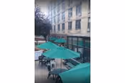 Outdoor patio area with several green umbrellas shading tables and chairs, adjacent to a multi-story building with multiple windows.