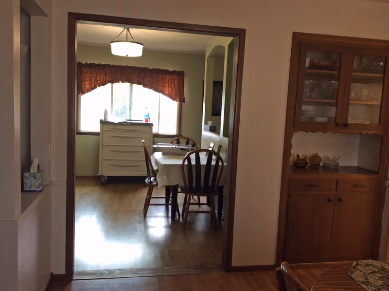 View through a doorway into a dining area with a table covered by a white tablecloth and four wooden chairs. A window with a red valance curtain is in the background, and a white cabinet with items on top is positioned under the window. To the right, there is a wooden china cabinet with glass doors displaying dishes and decorative items.