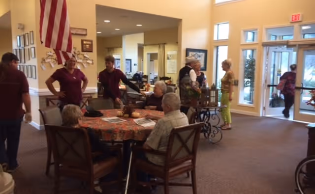 Interior common area of a senior living facility with residents and staff gathered around a decorated table near the entrance and large windows.