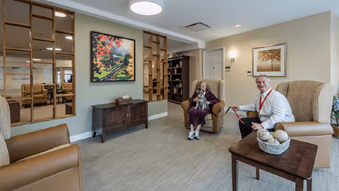A cozy living room area in a senior living facility with beige walls and carpeted floor. Two people are seated in armchairs; an elderly woman holding a small dog and a man holding the dog's leash. The room features a wooden sideboard with a colorful painting above it, a wooden coffee table with decorative items, and a large mirror on the wall reflecting part of the room.