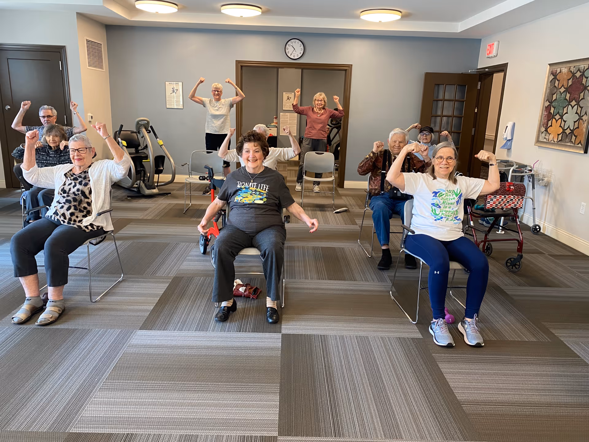 A group of elderly people sitting on chairs in a room, smiling and flexing their arms as if showing strength. The room has exercise equipment, a clock on the wall, and a colorful framed artwork. The floor is carpeted with a geometric pattern.