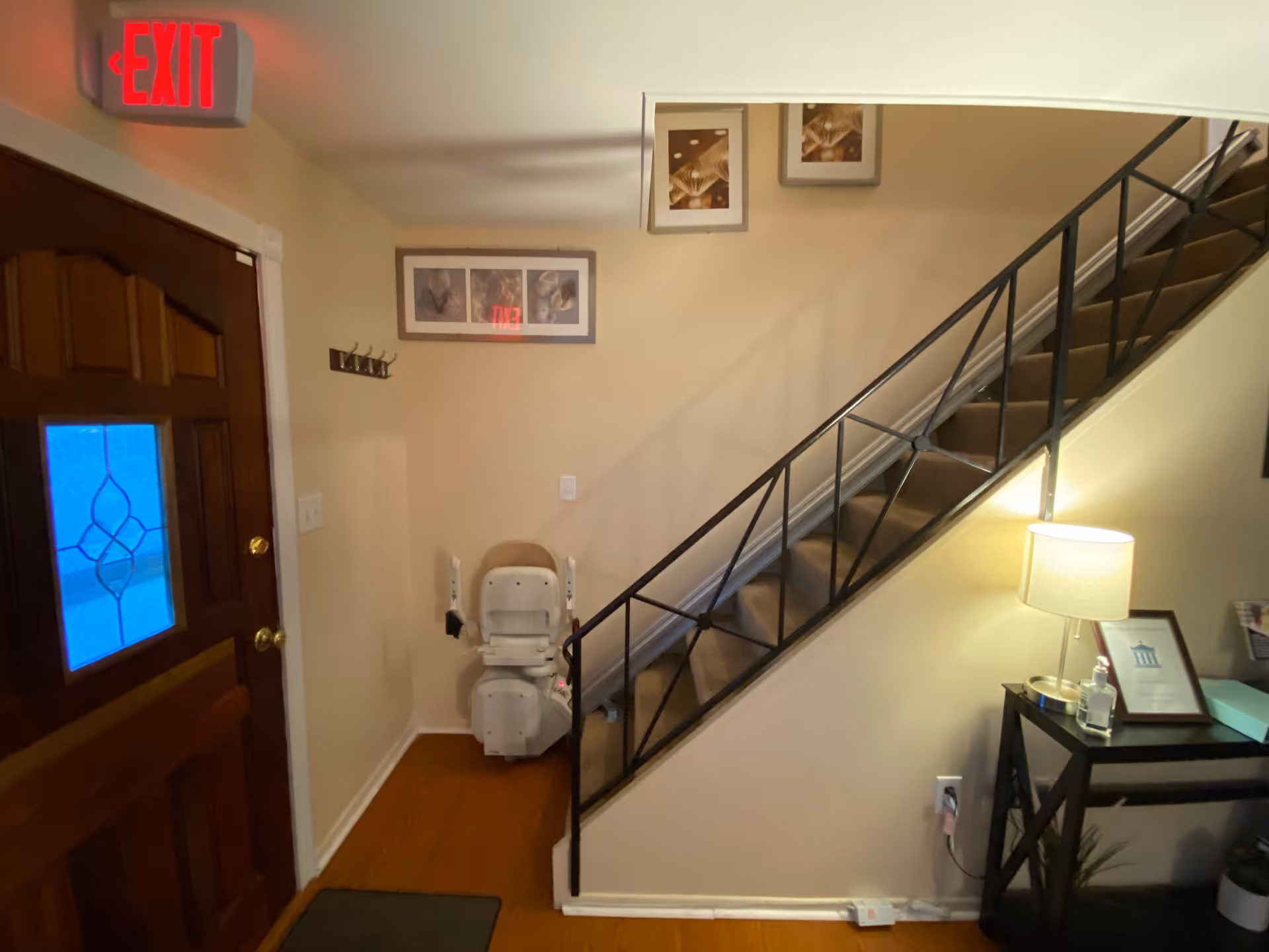 Interior view of a hallway with a wooden door on the left, an illuminated red exit sign above the door, a stairlift installed on a carpeted staircase with a black metal railing, and a small table with a lamp, hand sanitizer, and framed items on the right side.