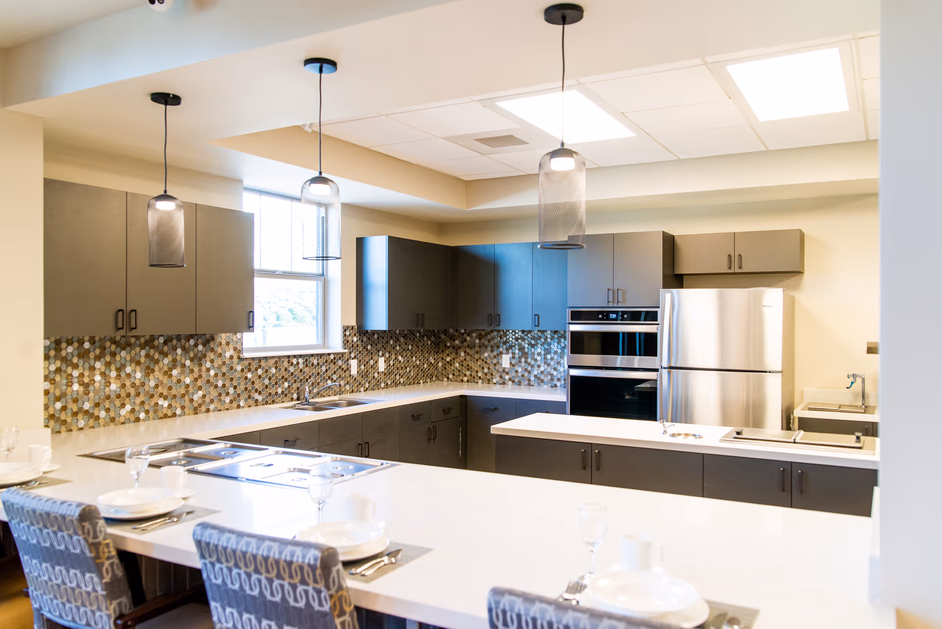Modern kitchen with dark cabinets, a mosaic tile backsplash, stainless steel refrigerator and oven, three pendant lights hanging from the ceiling, and a white countertop with place settings and patterned chairs.