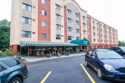 Exterior view of a multi-story senior living facility named The Harbor Court with a green awning over the entrance and several parked cars in front. The building has a beige and red facade with multiple windows and a covered porch area with rocking chairs and potted plants.