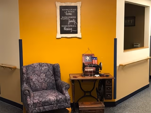 A cozy corner in a senior living facility featuring a patterned armchair next to a vintage sewing machine table with patriotic decorations. The wall behind is painted mustard yellow with a framed inspirational quote about home, family, and blessings.