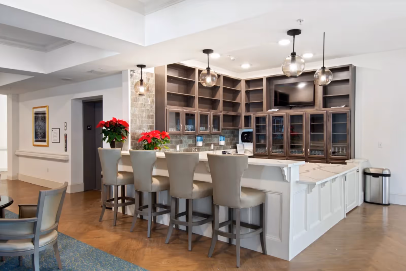 Interior view of a senior living facility lounge area featuring a white marble countertop bar with four beige cushioned bar stools. Behind the bar are wooden shelves and cabinets with glass doors, a mounted flat-screen TV, and three hanging pendant lights with glass globes. Two red poinsettia plants are placed on the bar counter. The floor is a combination of wood and blue carpet, and there is an elevator door visible in the background.