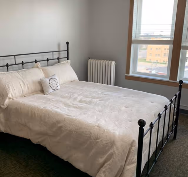 Bright bedroom with a black metal bed frame dressed in white linens beside a radiator and two windows.