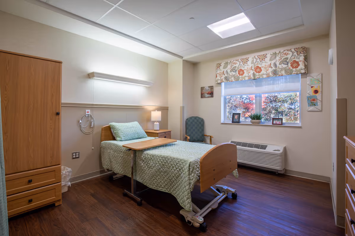A well-lit nursing facility bedroom with a single hospital-style bed covered in a green quilted bedspread. The room has wooden furniture including a wardrobe, nightstand with a lamp, and a small table on wheels positioned over the bed. There is a green upholstered chair near a window with floral valance and a white roller shade. The window sill holds framed photos and a potted plant. The floor is dark wood, and the walls are painted in neutral tones.