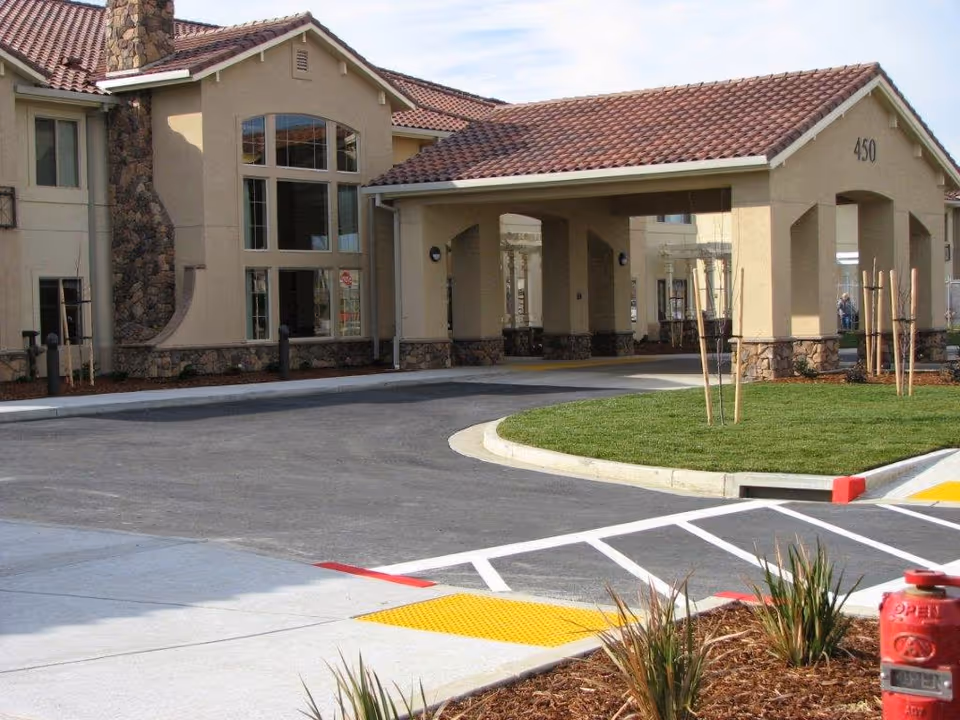 Exterior view of a senior living facility with beige walls, stone accents, and a red tiled roof. The building features large windows and a covered entrance with the number 450 displayed. There is a curved driveway, landscaped grass, and small plants in the foreground.