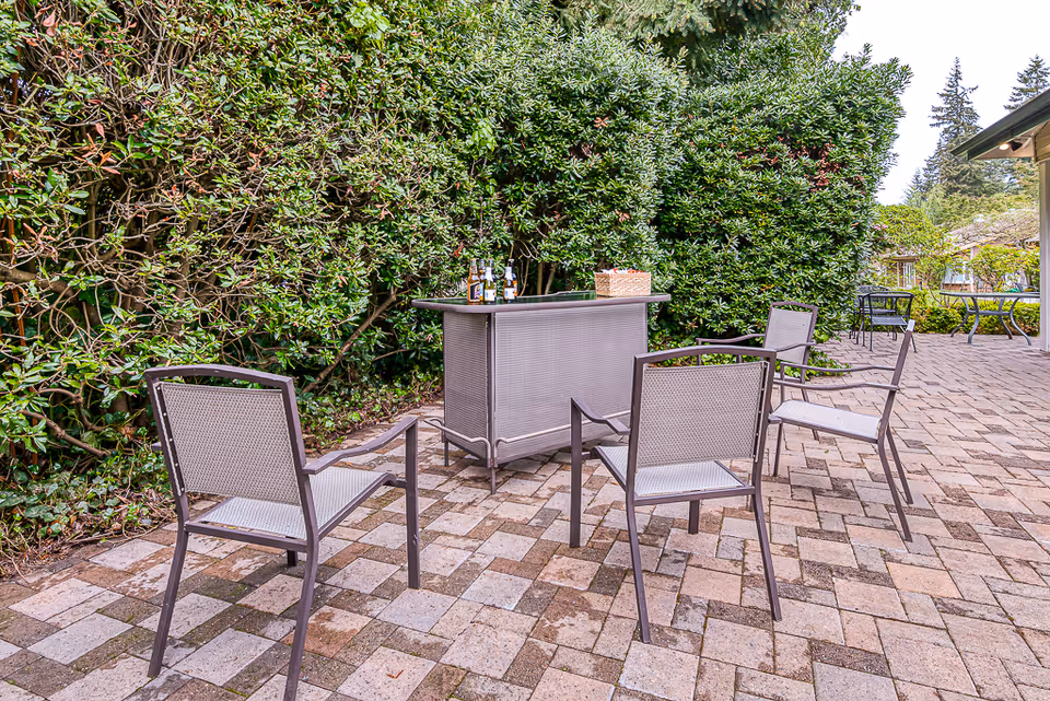 Outdoor patio area with four metal chairs arranged around a small bar table with bottles and a basket on top, surrounded by dense green bushes and trees.