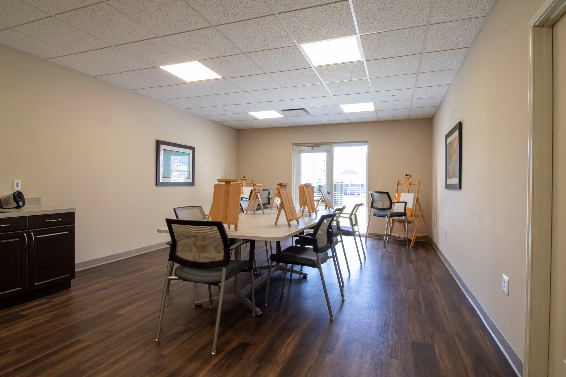 A well-lit room with a long table surrounded by chairs and several wooden easels placed on the table and around the room. The room has wood flooring, beige walls, a framed picture on the wall, and a door with windows letting in natural light.