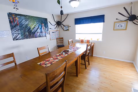 A dining room with a long wooden table and six wooden chairs. The table has a decorative runner with a fall leaf pattern and a few small items on it. The room has light-colored walls and a wooden floor. There is a window with a blue valance letting in natural light. Halloween decorations, including large black spiders, are attached to the walls.